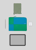 Layout piatto di elementi essenziali moderni: tablet, notebook con copertina a quadretti, M Patchwork Pouch Blue & Green, stilo e auricolari wireless su sfondo grigio.