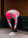 Personne portant un pull à rayures arc-en-ciel et une casquette rouge se penchant pour lacer ses chaussures en toile colorée dans une rue de la ville.