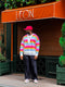 Un uomo con un maglione a righe colorate, sneakers e un cappello rosa si trova davanti a un ristorante chiuso chiamato The Smiles, con del verde intorno.