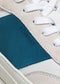Primer plano de una zapatilla baja beige con cordones blancos y etiqueta turquesa con la inscripción "V9 White & Petrol Blue".
