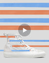 Phrase avec le nom du produit : une chaussure blanche en toile sur un fond rayé orange et bleu avec un bouton de lecture en surimpression, indiquant une nouvelle vidéo moyenne 1/5.